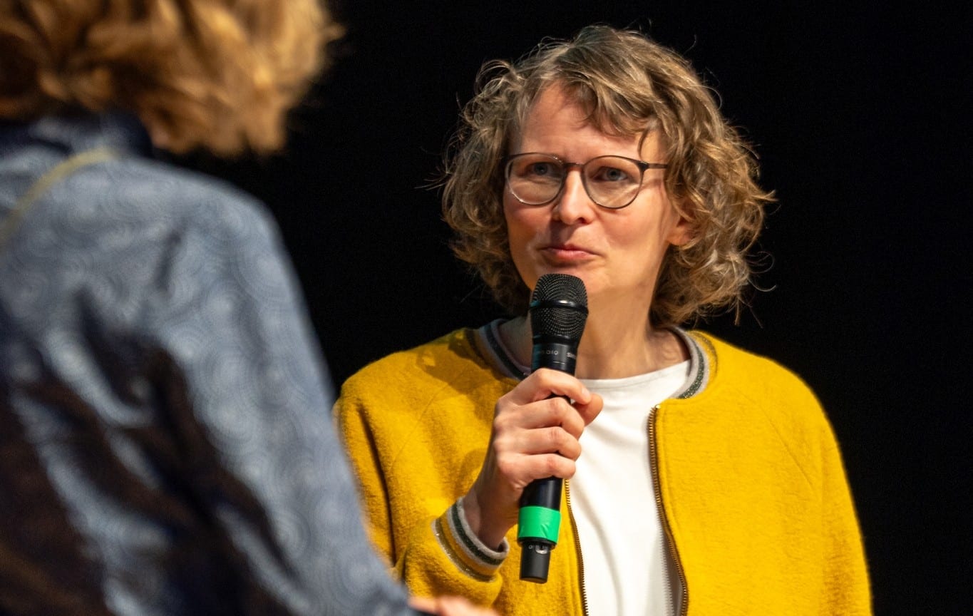 Eva in gelber Jacke spricht auf dem Podium ins Mikrofon, im Vordergrund eine Gesprächspartnerin von hinten