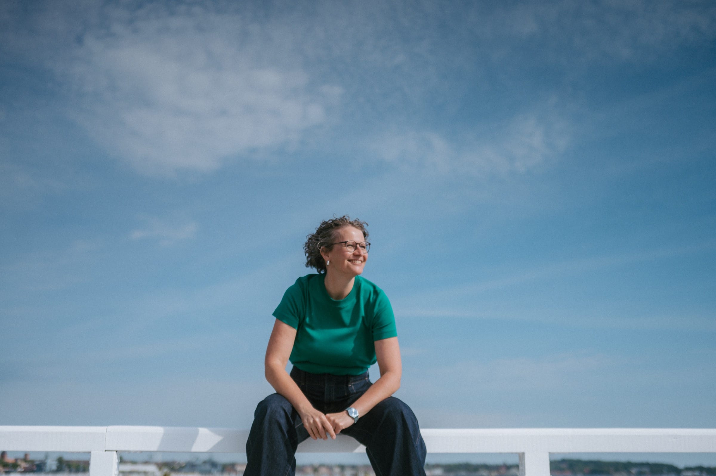 Eva sitzt lächelnd auf einem weißen Geländer am Wasser, über ihr weiter blauer Himmel