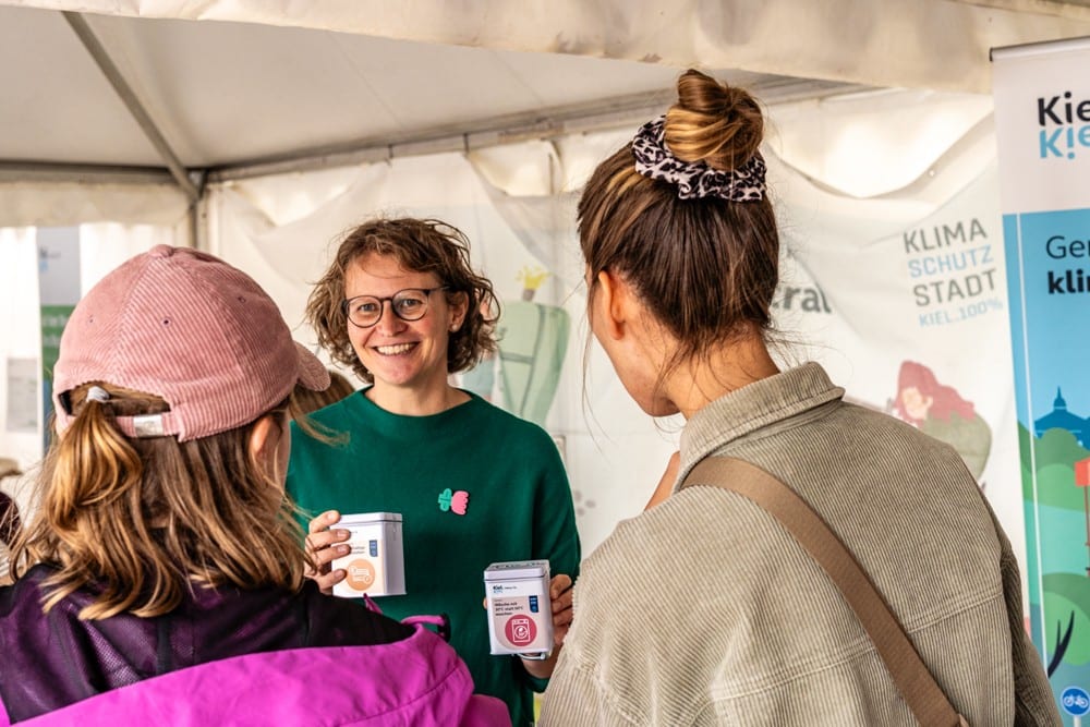 Eva lacht im Gespräch mit zwei Besucherinnen an einem Infostand und hält zwei kleine Dosen in den Händen